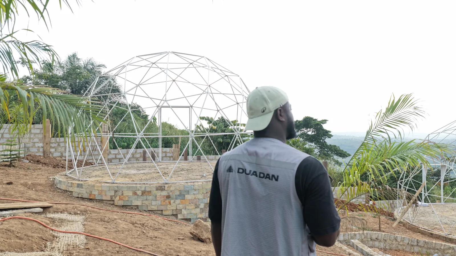 A geodesic dome frame standing on a circular stone-faced plinth cut into the hillside, with the valley beyond.